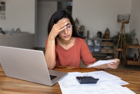 Stressed concerned young Hispanic woman working on financial documents, finding overspending in personal budget, holding head at home table with calculator, laptop and paper bills